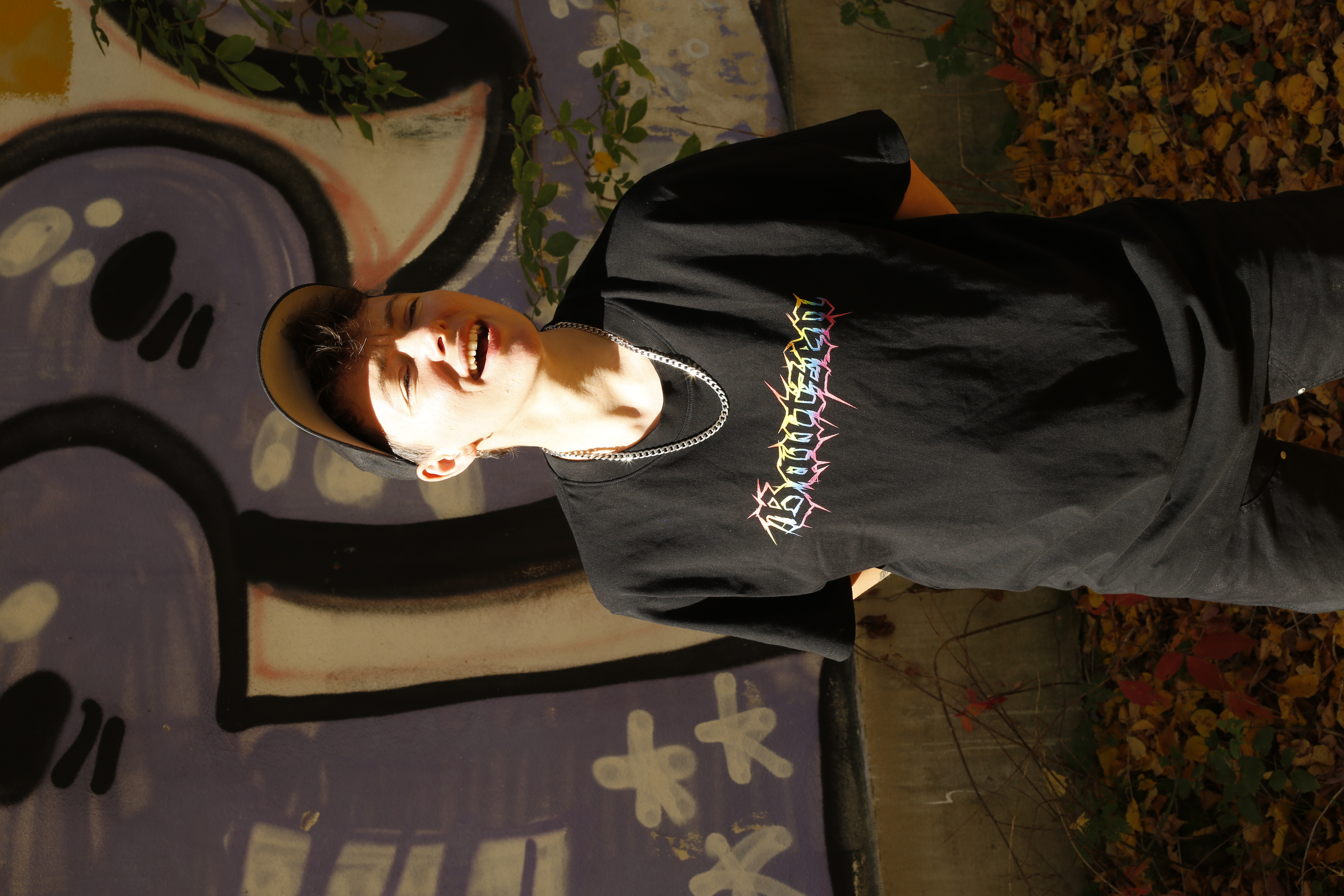 Mike steht draußen vor einer Mauer, die mit Grafitti besprüht ist. Mike trägt das schwarze CSD Bautzen Soli Shirt und eine schwarze Hose und eine schwarze Cappi. Die Sonne scheint Mike so sehr ins Gesicht, dass Mike lachen muss und die Augen zusammenkneift.