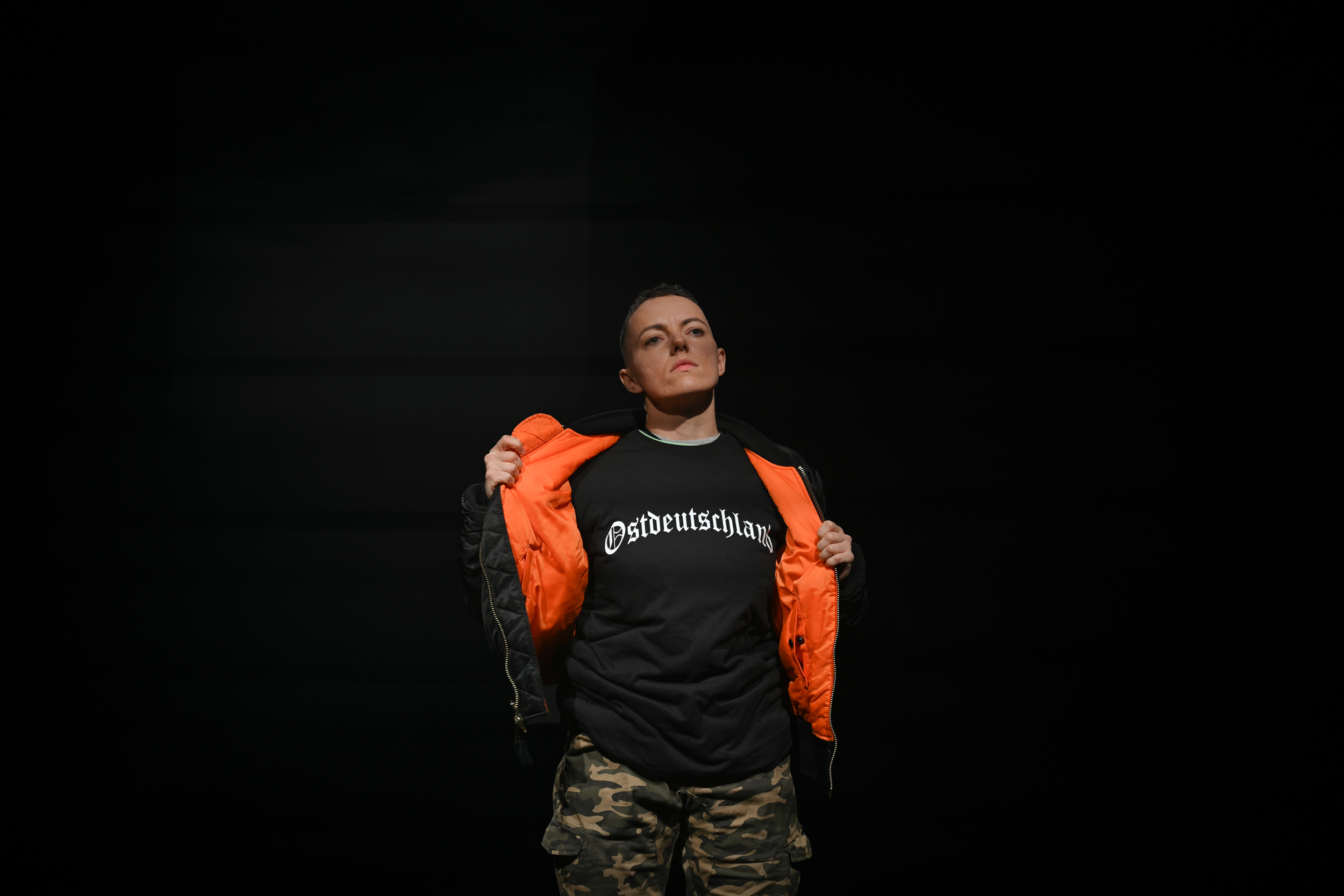 Mike öffnet die schwarze (von innen orangene) Bomberjacke. Darunter kommt ein schwarzes Tshirt mit der Aufschrift "Ostdeutschland" zum Vorschein. Mike hat sehr kurze Haare und guckt ernst.