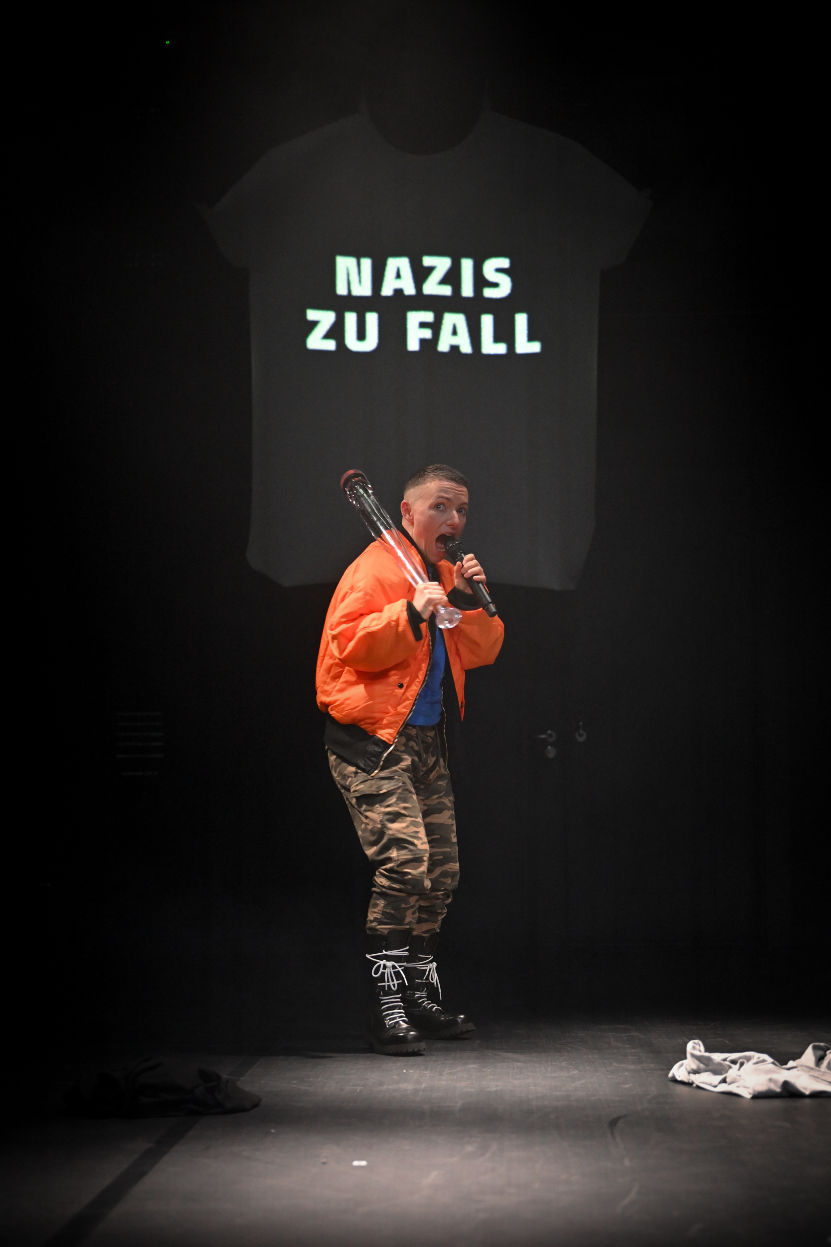 Mike steht auf der Bühne in orangener Bomberjacke, mit gläsernem Baseballschläger über der Schulter und rapt in ein Mikrofon. Auf der T-Shirt-förmigen Projektionsfläche dahinter stehen die Worte "Nazis zu Fall".