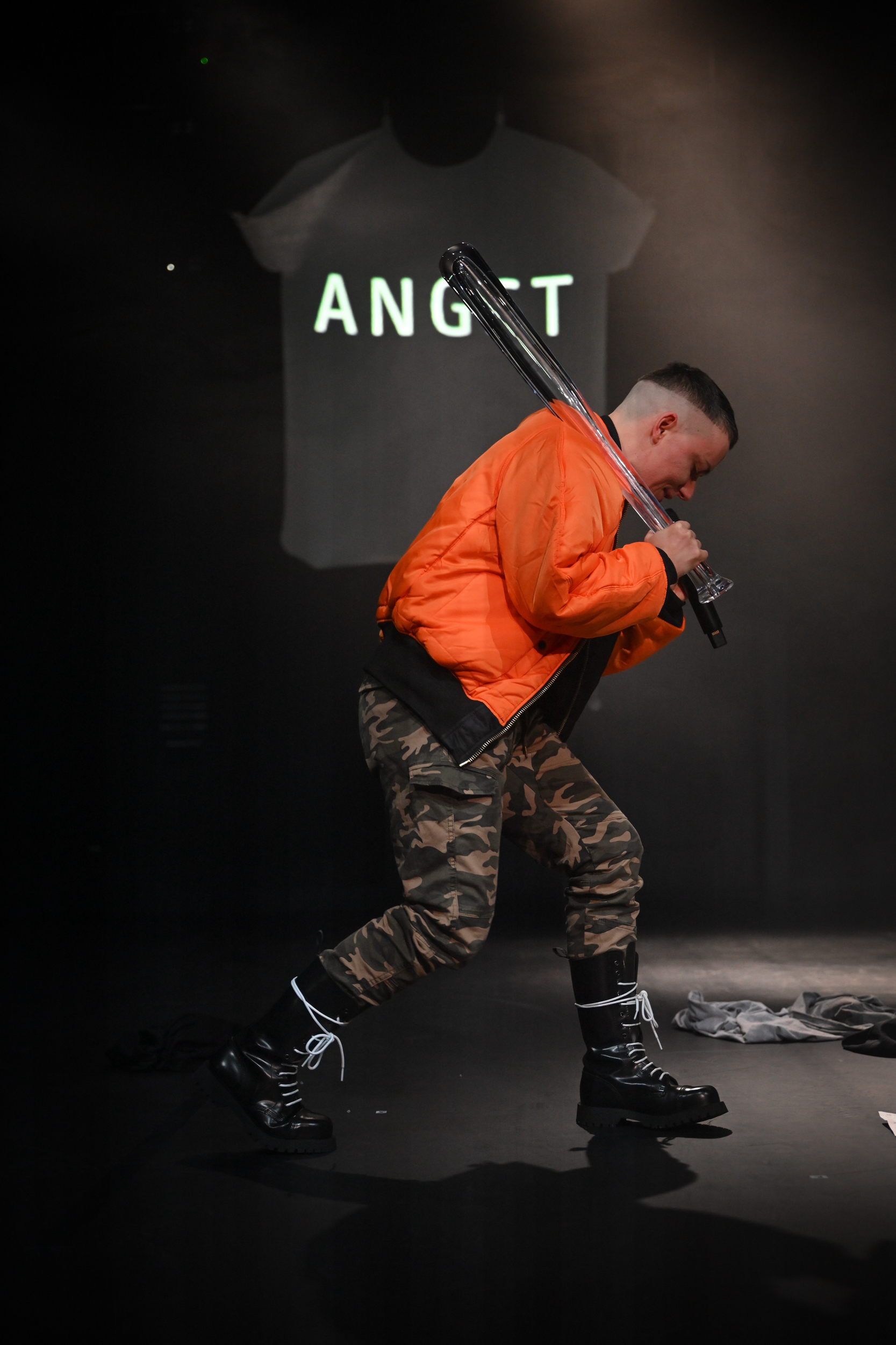 Mike steht seitlich zur Bühne in orangener Bomberjacke, mit gläsernem Baseballschläger über der Schulter und singt in ein Mikrofon. Auf der T-Shirt-förmigen Projektionsfläche dahinter steht das Wort Angst.