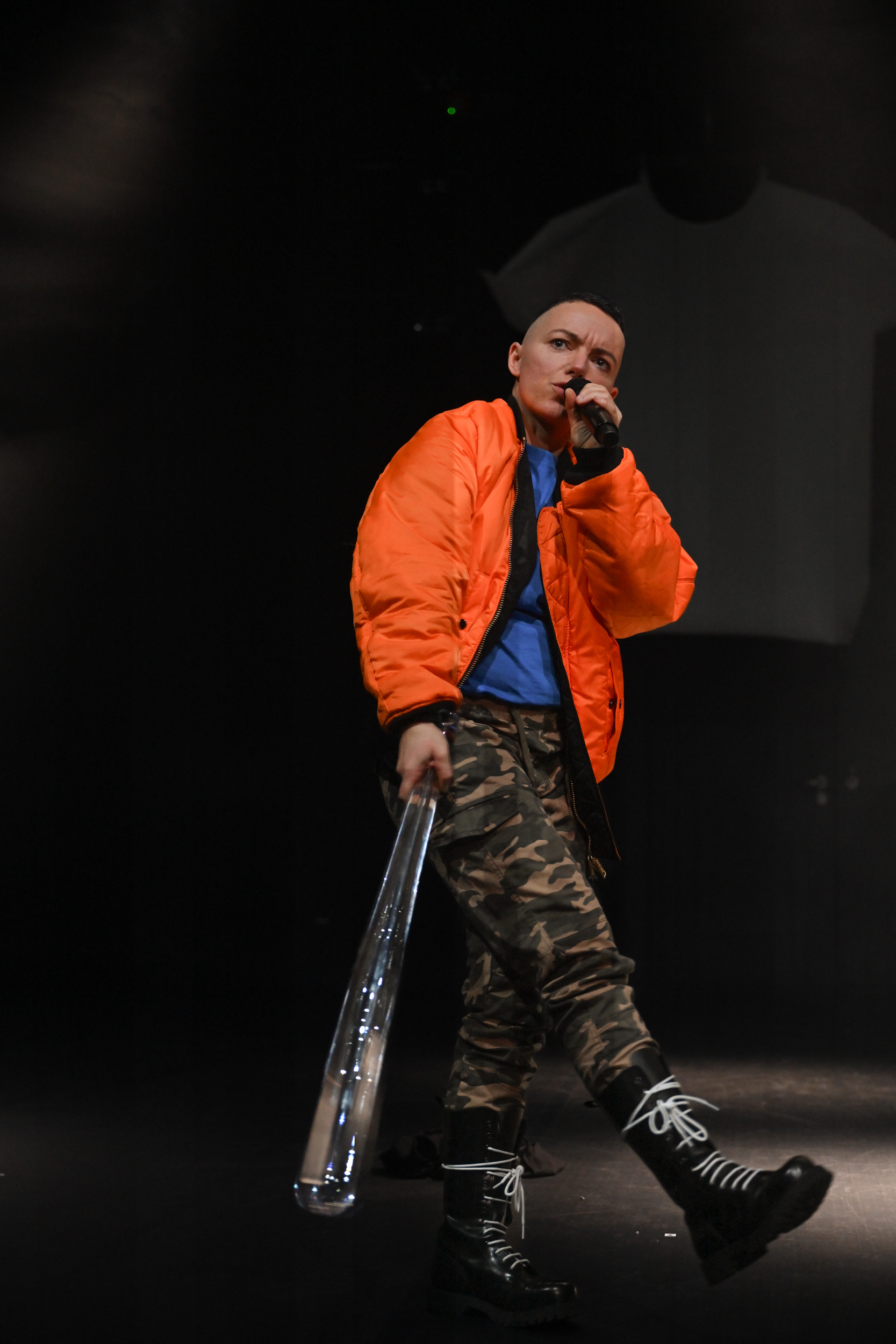 Mike läuft seitlich die Bühne entlang in orangener Bomberjacke und schwingt einen gläsernen Baseballschläger. Mike rapt in ein Mikrofon.