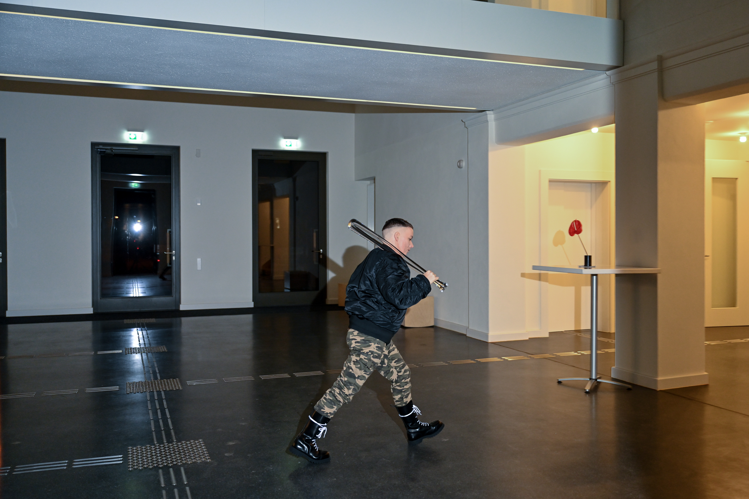 Maik läuft durch das Foyer des Ostflügels in Hellerau. Maik trägt Bomberjacke, Armyhosen und einen gläsernen Baseballschläger über der Schulter.