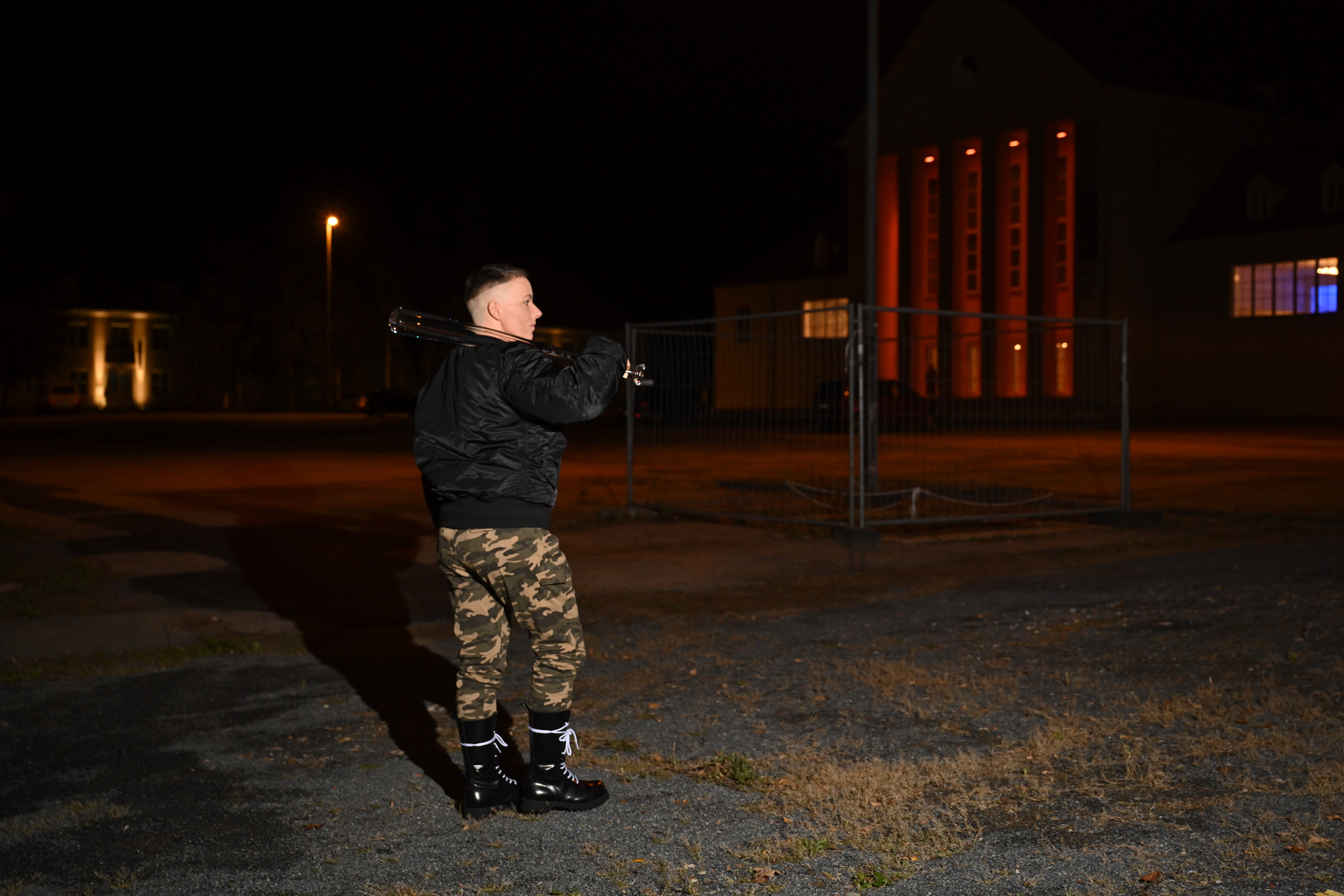 Maik steht auf dem Vorplatz vor dem Festspielhaus Hellerau. Es ist Nacht. Maik trägt Bomberjacke, Armyhosen und einen gläsernen Baseballschläger über der Schulter.