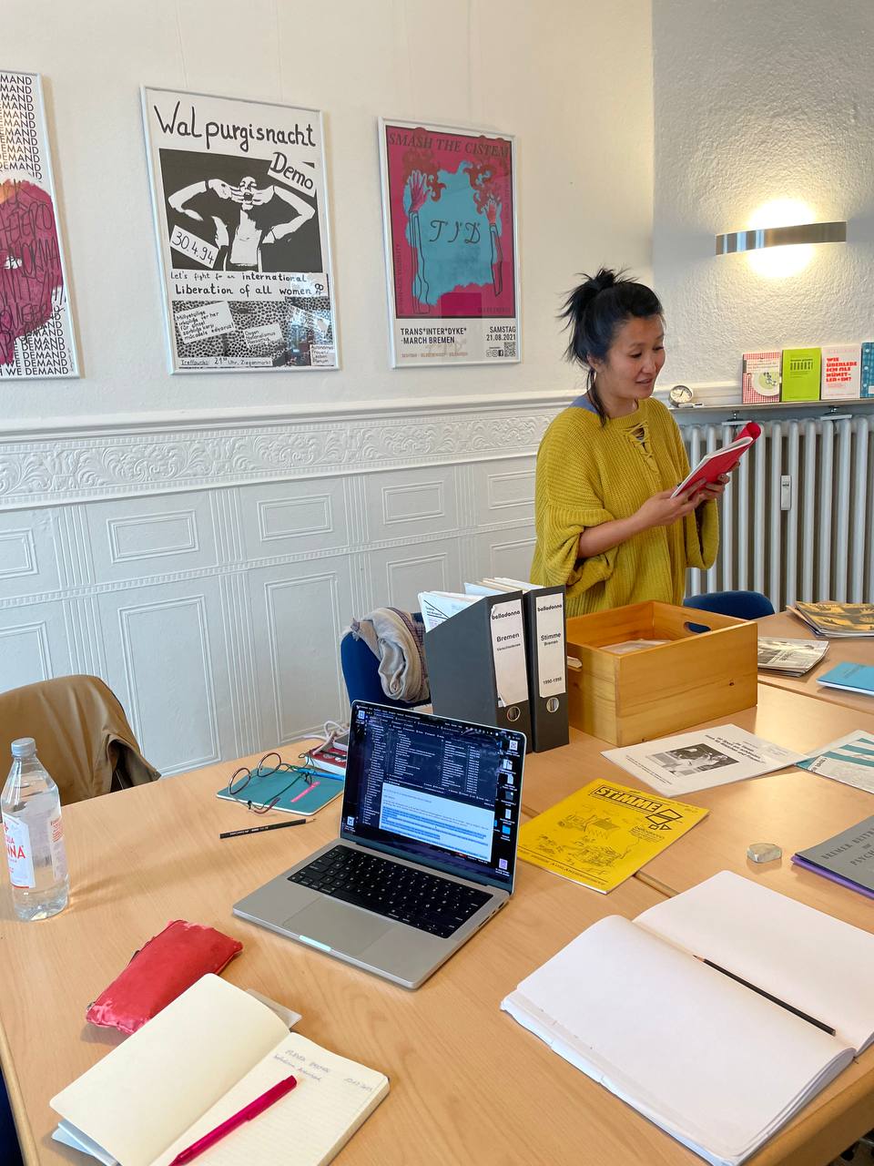 Melmun steht am Tisch im Belladonna-Archiv Bremen und liest aus einem zine der 90'er Jahre vor. Hinter Melmun an der Wand hängen Plakate von feministischen Aktionen und Protesten. Auf dem Tisch vor Melmun liegen ein aufgeklappter Laptop und ein Notizbuch sowie weitere Ordner mit Recherchematerialien.