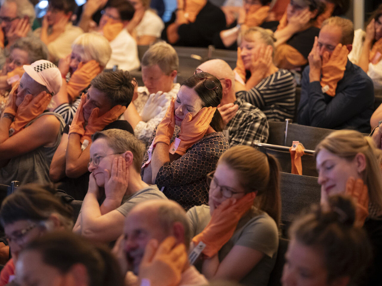 Das Publikum im Saal der historischen Bauhausbühne Dessau sitzt in Reihen hintereinander. Alle haben sich Anziehbare (orangene Handschuhartige Stoffstücke) übergestreift und folgen den choreografischen Anweisungen von Mike. Auf dem Bild halten die meisten gerade ihr Gesicht mit beiden Händen umschlossen.