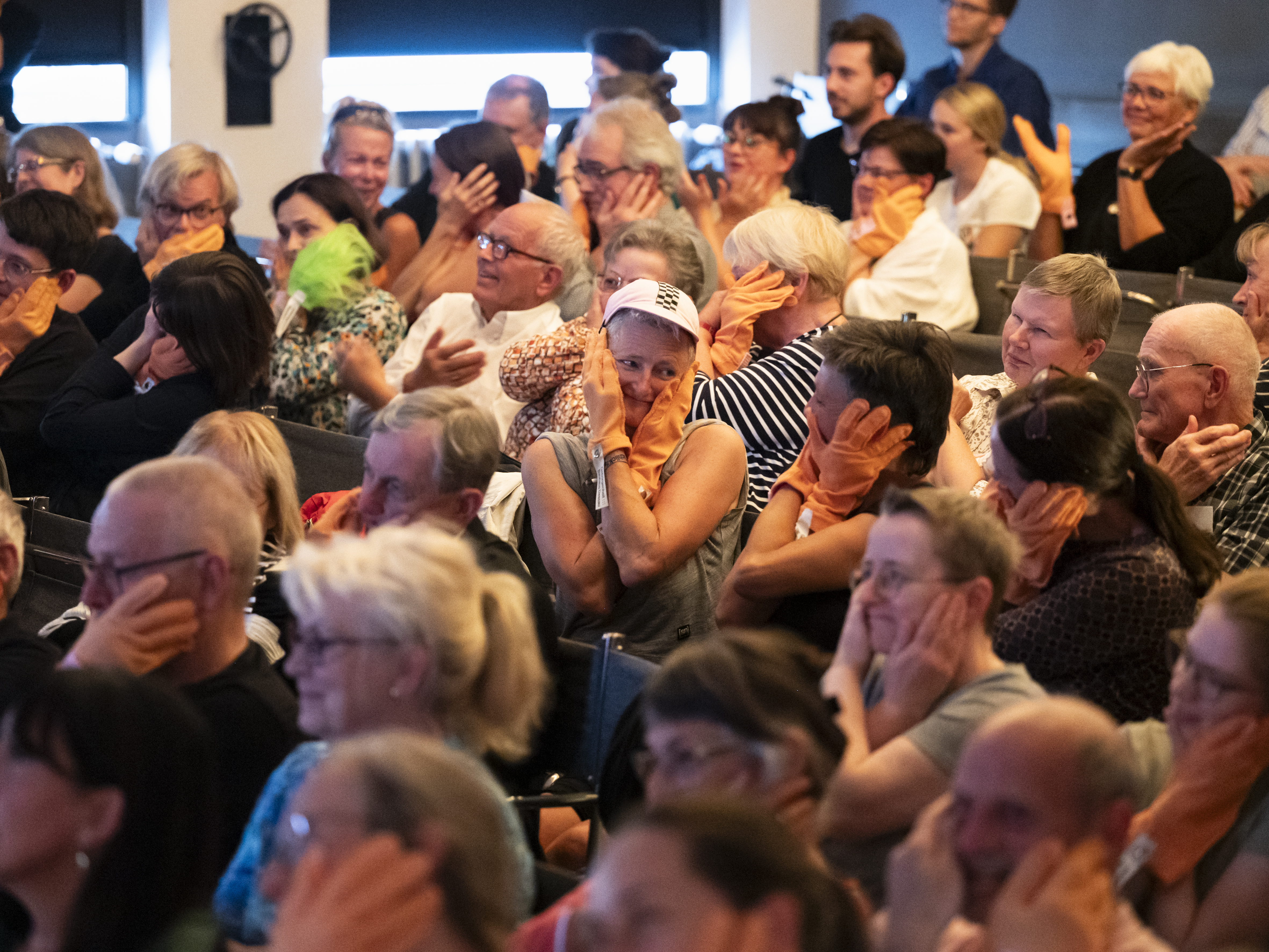 Das Publikum im Saal der historischen Bauhausbühne Dessau sitzt in Reihen hintereinander. Alle haben sich Anziehbare (orangene Handschuhartige Stoffstücke) übergestreift und folgen den choreografischen Anweisungen von Mike. Auf dem Bild halten die meisten gerade ihr Gesicht mit beiden Händen umschlossen. Zwei Personen in der Bildmitte schauen einander belustigt an.