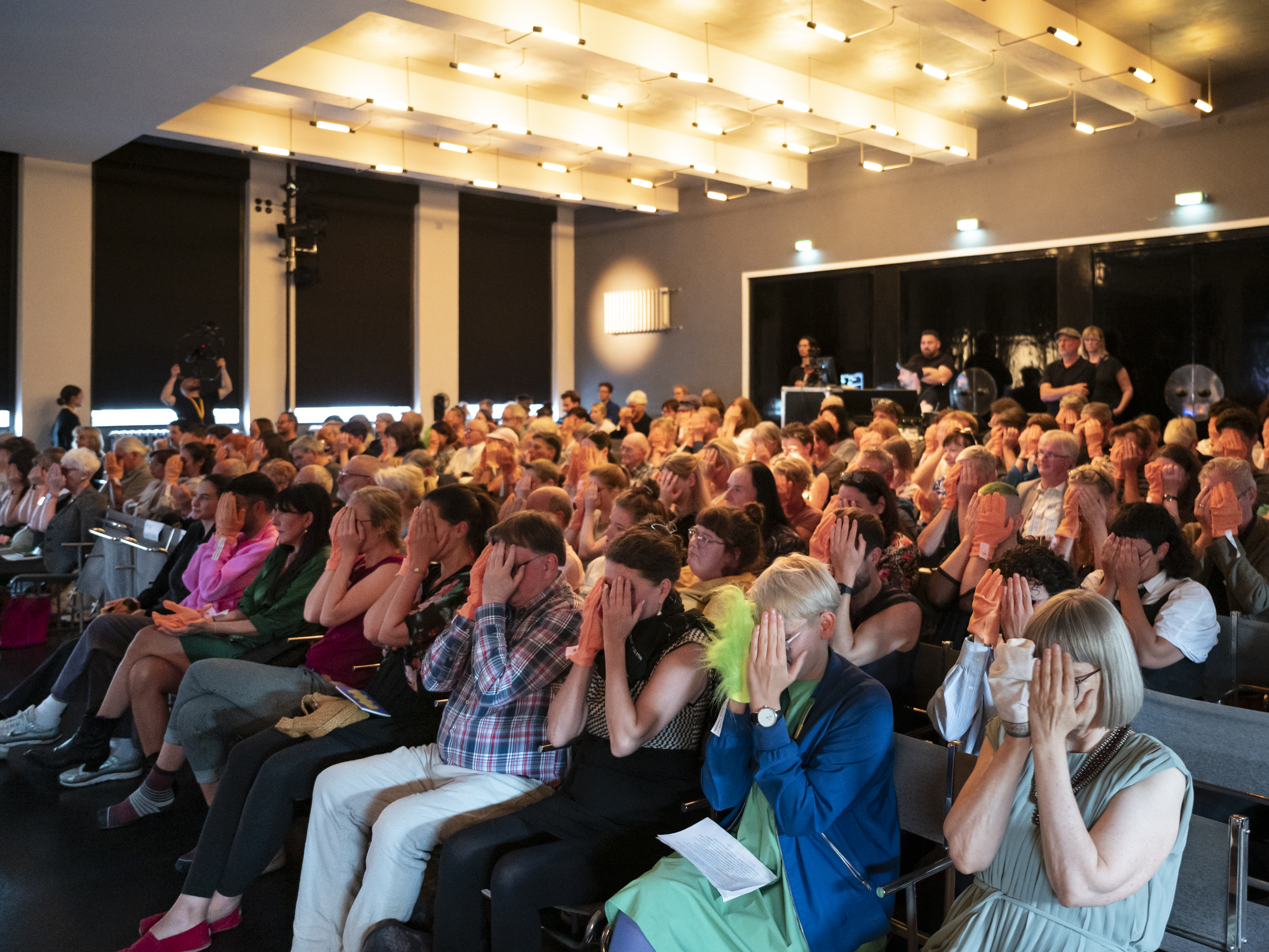 Das Publikum im Saal der historischen Bauhausbühne Dessau sitzt in Reihen hintereinander. Alle haben sich Anziehbare (orangene Handschuhartige Stoffstücke) übergestreift und folgen den choreografischen Anweisungen von Mike. Auf dem Bild halten die meisten gerade beide Hände vor ihr Gesicht. Auf dem Fotoausschnitt ist der gesamte Publikumsraum der Bauhausbühne abgebildet.