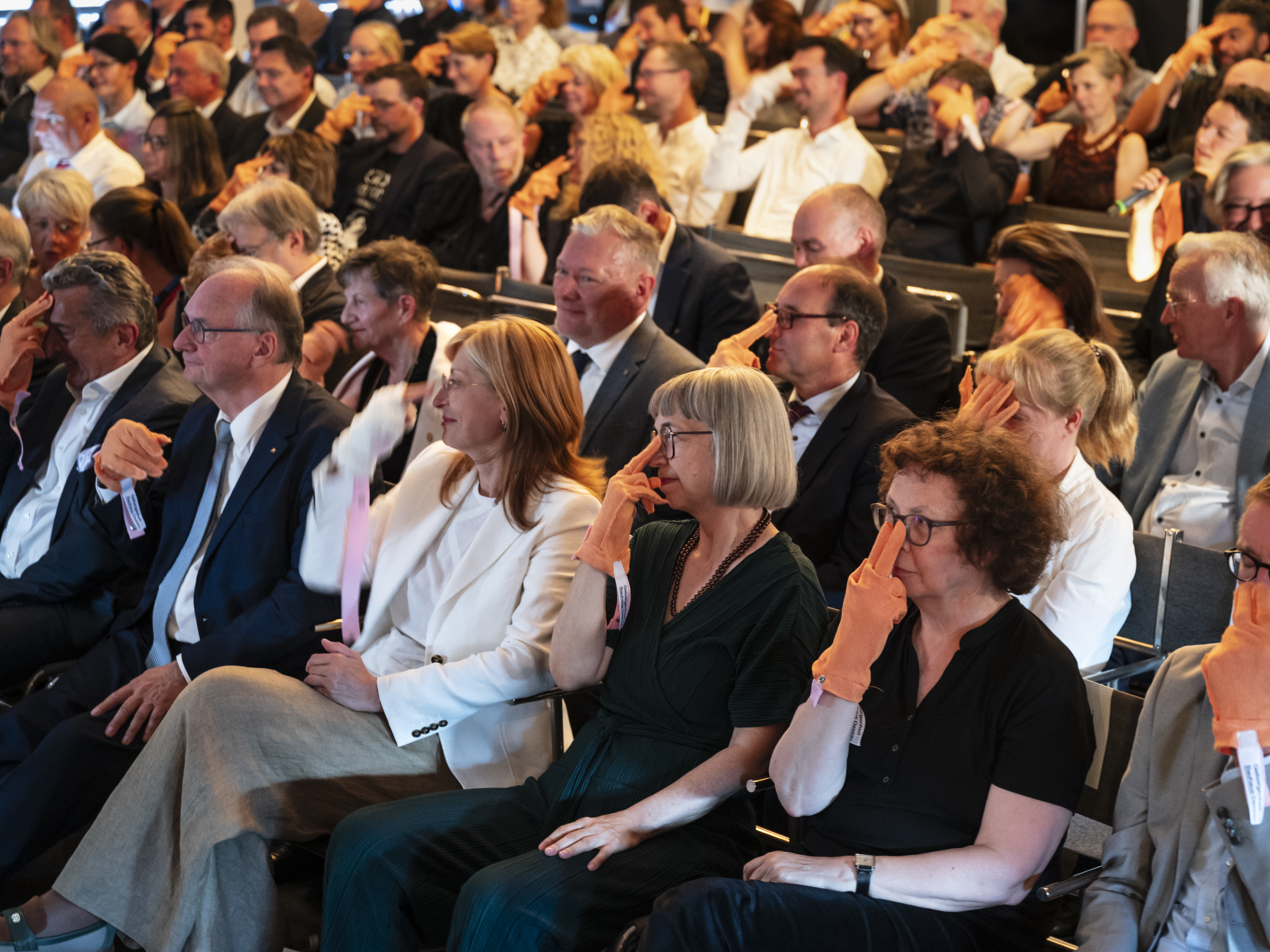 Das Publikum im Saal der historischen Bauhausbühne Dessau sitzt in Reihen hintereinander. Alle haben sich Anziehbare (orangene Handschuhartige Stoffstücke) übergestreift und folgen den choreografischen Anweisungen von Mike. Hier legen die meisten gerade zwei Finger auf ihren Nasenrücken.