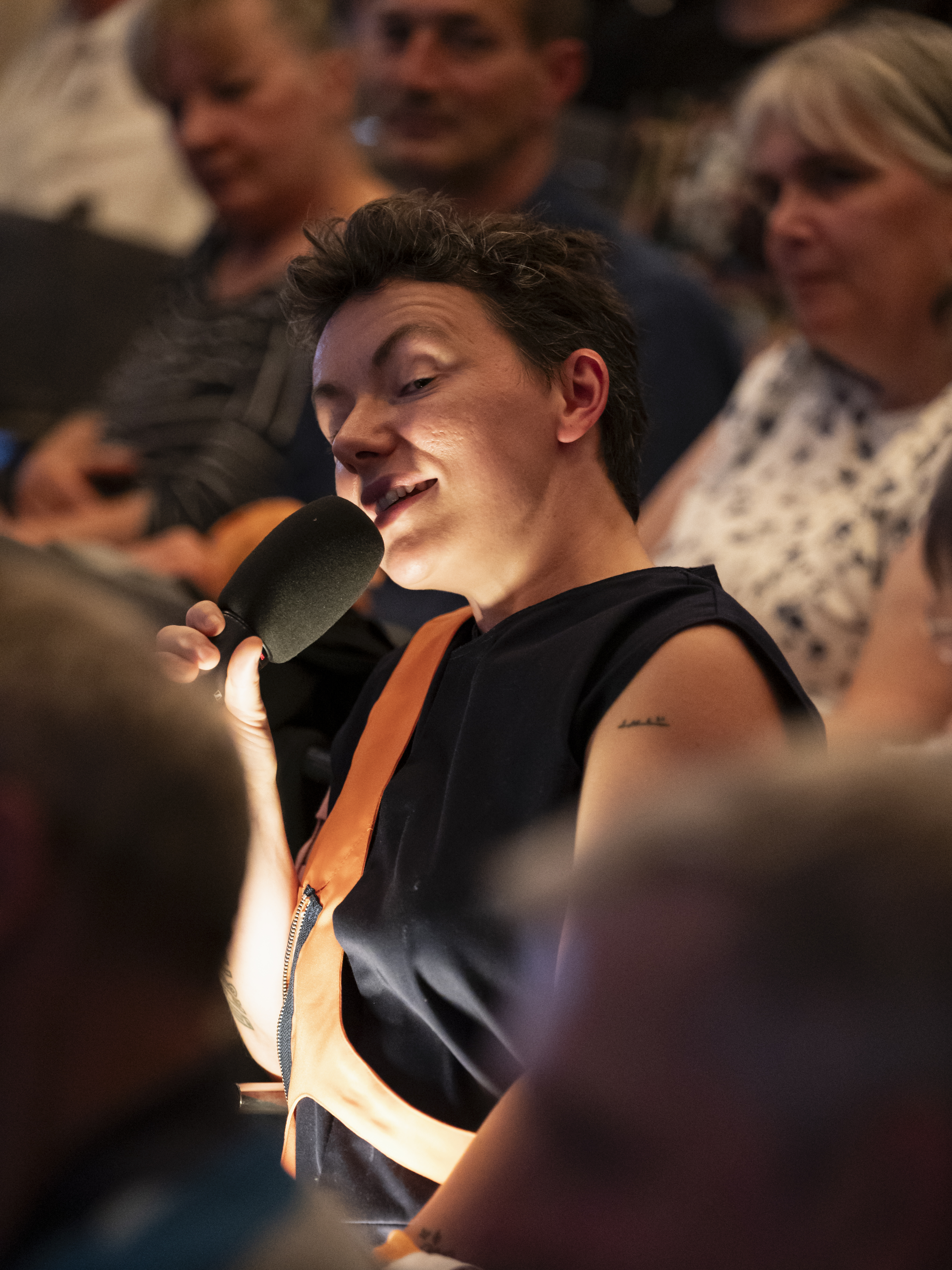 Mike sitzt in den Reihen zwischen dem Publikum, spricht in ein Mikrofon und wird von unten beleuchtet. Mike hat dabei einen schelmischen Gesichtsausdruck.