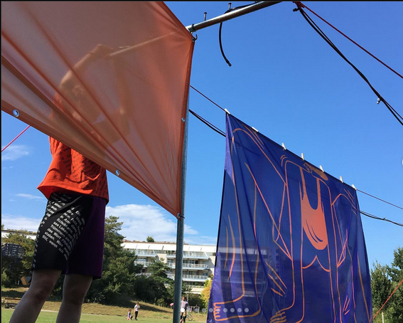 Eine Person in einer schwarzen Kostümhose und orangenem Kostümshirt steht hinter einem orangenen Sonnensegel. Neben der Person weht ein lila Sonnensegel mit orangener Zeichnung. Die Segel sind an einer Wäscheständerkonstruktion befestigt, die im öffentlichen Stadtpark St. Johann von Basel steht.